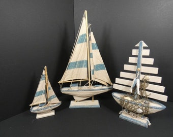 May include: Three decorative wooden sailboat models with blue and white striped sails. The largest sailboat is in the center, the smallest is on the left, and the middle-sized sailboat is on the right. The sailboats are on white bases.