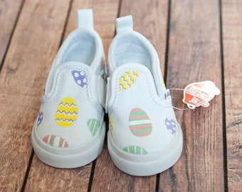 May include: A pair of white canvas slip-on shoes with an Easter egg design. The eggs are painted in various colors, including yellow, purple, green, and pink.