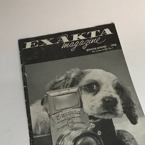 May include: A black and white photo of a puppy holding a black and silver Exakta camera. The photo is from the Exakta magazine, Winter-Spring 1958. The magazine cover also includes a yellow advertisement for Exakta publications.