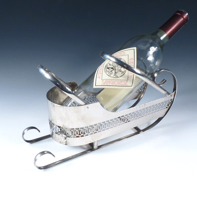 May include: A silver-toned metal wine bottle holder shaped like a sleigh with curved runners and a decorative openwork band along the sides. A clear glass wine bottle with a label reading Sangiovese is positioned inside the metal cradle.