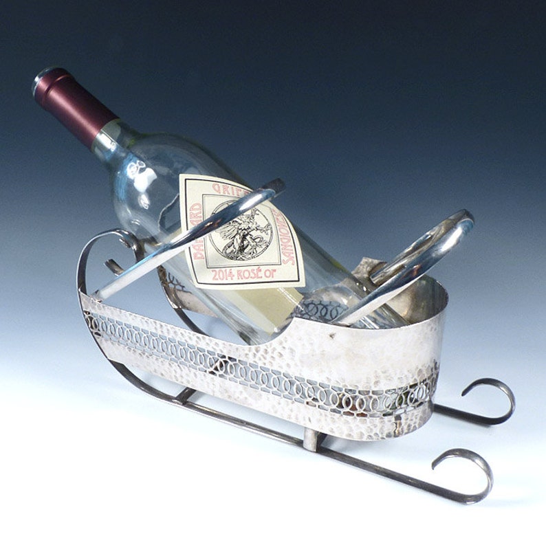 May include: A silver-toned metal wine bottle holder shaped like a sleigh with a hammered texture and an openwork oval pattern along the sides. A clear glass wine bottle with a label reading 2014 ROSE OR is resting inside the metal sleigh frame.