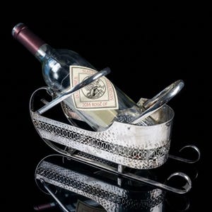 May include: A silver-toned metal wine bottle holder shaped like a sleigh with a hammered texture and a decorative circular link pattern along the sides. A clear glass wine bottle with a label reading 2014 ROOZ OF is resting inside the holder.