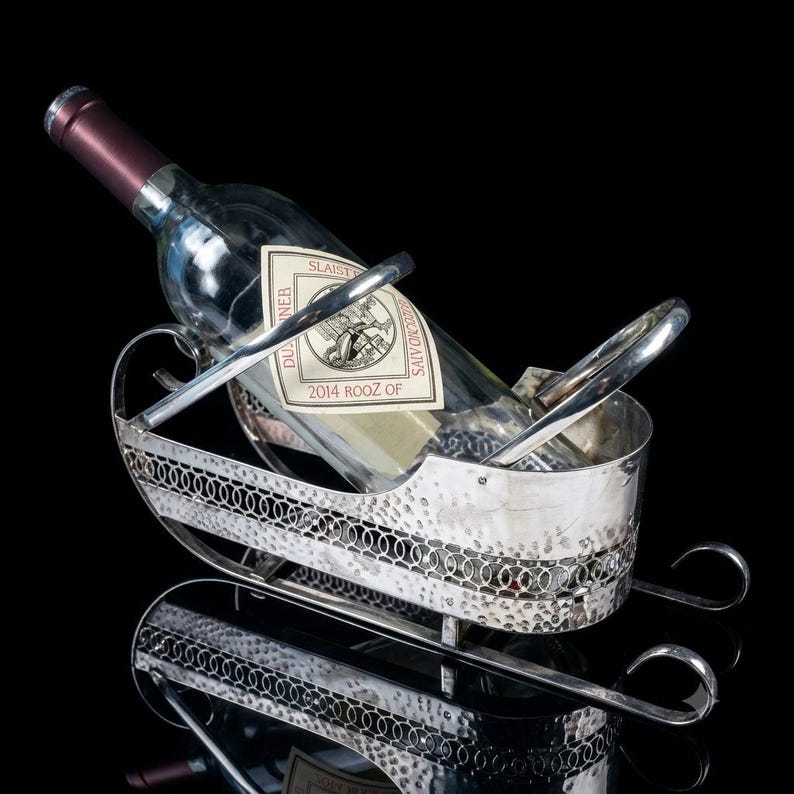 May include: A silver-toned metal wine bottle holder shaped like a sleigh with a hammered texture and a decorative circular link pattern along the sides. A clear glass wine bottle with a label reading 2014 ROOZ OF is resting inside the holder.
