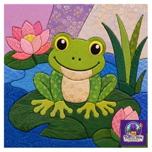 May include: A fabric applique wall hanging featuring a green frog sitting on a large lily pad in a blue pond. The scene includes pink lotus flowers, green reeds, and a purple and white background. A logo for Turtle Sign Designs is in the corner.