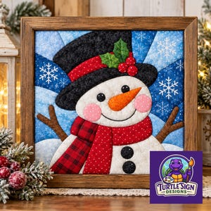 May include: A square framed fabric wall art piece featuring a quilted snowman with a black top hat, a red plaid scarf, and an orange nose. The background is a blue starburst pattern with white snowflakes. The snowman has pink cheeks and black buttons.