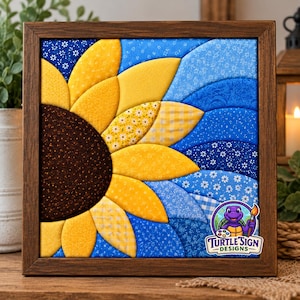 May include: A square fabric wall art piece in a dark wood frame featuring a quilted sunflower design. The sunflower has a dark brown center and yellow petals with various patterns. The background consists of layered blue fabric waves.