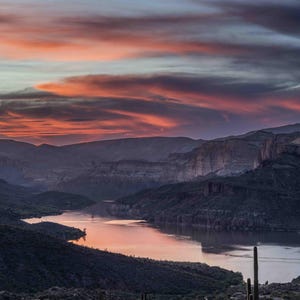 May include: A scenic landscape photograph of a river winding through a canyon at sunset. The sky is filled with vibrant orange and purple hues, reflecting in the water. Cacti are visible in the foreground.