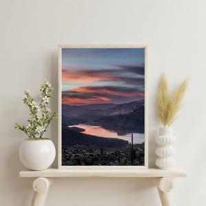 Könnte beinhalten: Gerahmter Kunstdruck mit einem Sonnenuntergang über einer Bergkette und einem See. Der Himmel zeigt Farbtöne von Orange, Rosa und Blau. Der Druck befindet sich in einem hellen Holzrahmen und erzeugt eine ruhige Szene.