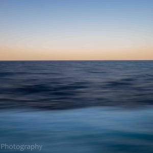 May include: An abstract seascape photograph with a colour gradient. The image shows a transition from a light blue sky to a golden horizon, with dark blue ocean waves in the foreground. The water appears blurred, suggesting movement.