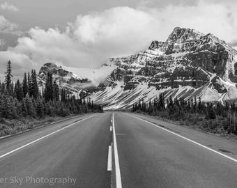May include: Black and white photograph of a mountain road leading towards snow-capped mountains under a cloudy sky. Tall evergreen trees line the sides of the road. The road has white lane markings.