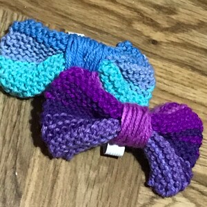 May include: Two knitted hair bows in shades of purple, blue, and teal. The bows are tied together with a purple yarn.