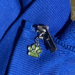 Op de afbeelding: Een blauwe rits met een zilveren metalen rits schuif en een groene glitter-pootafdruk charme.