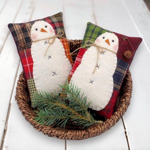 May include: Two snowman-shaped pillows with white felt bodies, orange beaks, and black cross stitch details. They rest in a woven basket with a sprig of evergreen. The pillows have plaid fabric backgrounds in red, green, and brown.