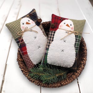 May include: Two snowman-shaped decorative pillows rest in a woven basket. The snowmen have white bodies, orange noses, and black cross stitch eyes. The pillows have plaid and checkered patterns in red, green, and navy blue.