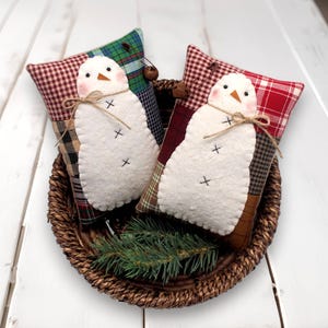 May include: Two snowman-shaped decorative pillows with white felt bodies, black cross stitch details, and carrot noses. They rest on patchwork pillows in a woven basket with a sprig of evergreen. The pillows are tied with twine.