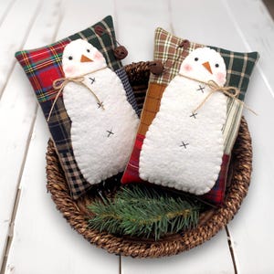 May include: Two decorative snowman pillows with white bodies, black cross stitch details, and carrot noses rest on plaid fabric pillows. The pillows are in a woven basket with a sprig of evergreen.