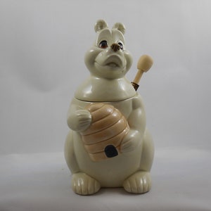 May include: A ceramic honey pot shaped like a bear holding a beehive. The bear is white with brown eyes and a wooden honey dipper is attached to the top of the pot.