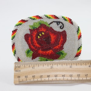 May include: A beaded oval-shaped pin with a red rose design. The rose is surrounded by white beads and a border of red, yellow, and green beads.