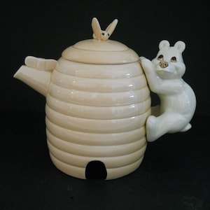 May include: Cream-colored ceramic teapot shaped like a beehive, with a bear climbing the side. The lid has a small bee figurine. The teapot has a spout and a handle. The beehive design has horizontal ridges.
