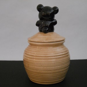 May include: A ceramic jar with a lid shaped like a black bear. The jar is tan with horizontal lines and a speckled texture.