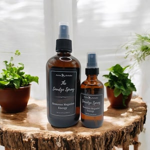 May include: Two brown glass bottles with spray tops. The larger bottle is labeled "The Smudge Spray" and the smaller bottle is labeled "Smudge Spray". Both bottles say "Removes Negative Energy".