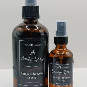 May include: Two brown glass bottles with black spray tops and labels. The larger bottle is labeled "The Smudge Spray Removes Negative Energy Net wt. 8 fl oz/236 ml" and the smaller bottle is labeled "Smudge Spray Removes Negative Energy Net Wt. 2 oz/60 ml". Both bottles have a white bird logo with the text "Herban Herbalist".