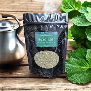 Puede incluir: Una bolsa negra con una etiqueta blanca que dice "Herbalist Head Ease Headache Relief Tea". La bolsa está sobre una superficie de madera con hojas verdes.
