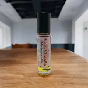 Puede incluir: Un frasco de vidrio transparente con tapa negra y una etiqueta roja y blanca que dice "ROOT CHAKRA Chakra Balancing Roll On Herbalist".
