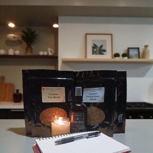 Könnte beinhalten: Eine brennende Kerze, ein Notizbuch mit Stift und mehrere schwarze Beutel mit Tee- und Bademischungen sind auf einer Arbeitsplatte angeordnet. Eine Sprühflasche von "The Smudge Spring" ist ebenfalls vorhanden. Die Szene deutet auf Kräuterwellness und Entspannung hin.