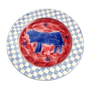 May include: A round ceramic plate featuring a blue and white checkered border. The center is red with a hand-painted blue cow silhouette surrounded by small blue stars and red leaf-like brushstrokes, creating a decorative dining piece.