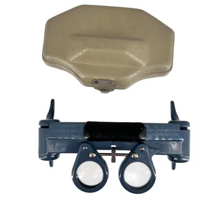 May include: A blue Abrams 2-4 stereoscope model CB-4 with two circular lenses, a central adjustment knob, and a black forehead rest. The device includes a textured beige protective case with a metal snap closure for storage and transport.