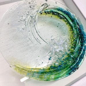May include: A round glass plate with a swirling design in shades of green and blue. The plate has a textured surface with small bubbles and flecks of colour.