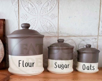 May include: Three ceramic canisters with lids, labeled "Flour", "Sugar", and "Oats". The canisters are brown with a white band around the middle. They are sitting on a wooden surface.
