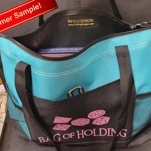 May include: A turquoise blue tote bag with black straps and a black mesh pocket. The bag has a zipper closure and a pink embroidered design that says "Bag of Holding" with dice imagery. The bag is sitting on a black chair.