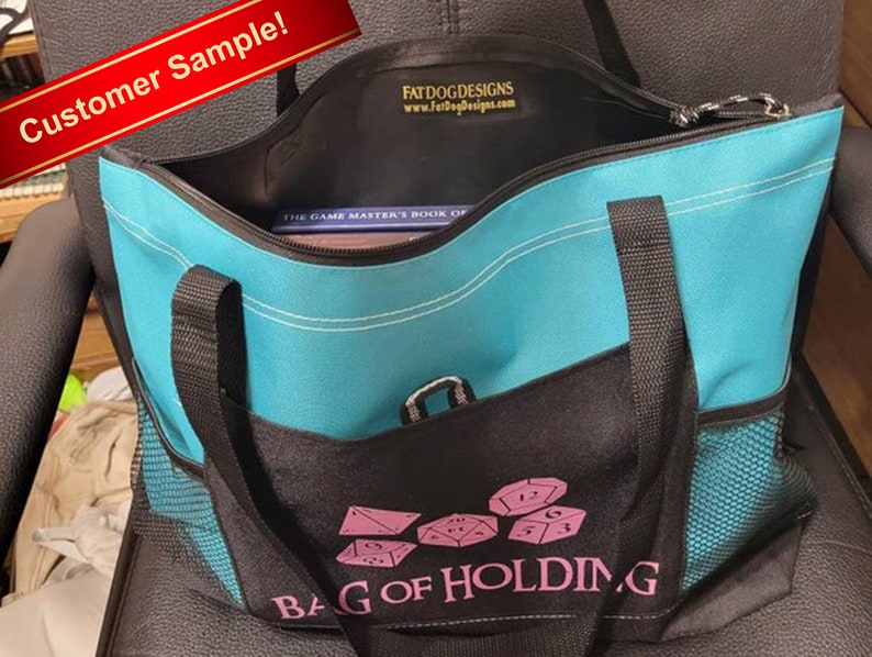 May include: A turquoise blue tote bag with black straps and a black mesh pocket. The bag has a zipper closure and a pink embroidered design that says "Bag of Holding" with dice imagery. The bag is sitting on a black chair.