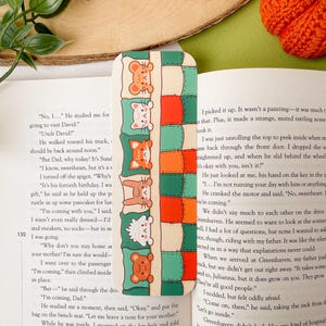 May include: A colorful bookmark featuring a repeating pattern of cartoon animals on pillows. The bookmark has a cream background with green, orange, and red accents. The animals include a bear, fox, cat, and bunny.