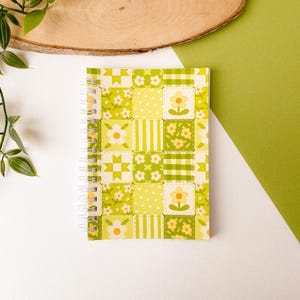 Könnte beinhalten: Ein spiralgebundenes Notizbuch mit einem Patchwork-Design mit gelben und grünen Quadraten. Die Quadrate sind mit floralen und geometrischen Mustern verziert. Das Notizbuch hat eine weiße Spiralbindung.