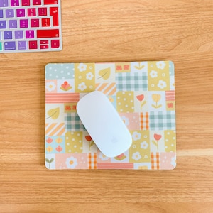 May include: A rectangular mouse pad featuring a patchwork design of yellow, light green, and orange squares with patterns of flowers, leaves, and gingham checks. A smooth, white wireless computer mouse rests on top of the patterned surface.