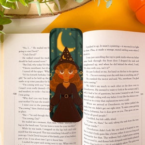 Puede incluir: Un marcapáginas con una ilustración de dibujos animados de una bruja con cabello castaño que lleva un sombrero puntiagudo negro y un vestido negro. La bruja está de pie en un bosque con una luna creciente en el fondo. El marcapáginas está hecho de un material resistente y es perfecto para marcar tu lugar en un libro.