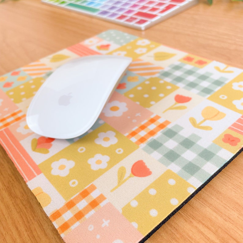 May include: A square mouse pad featuring a patchwork design of yellow, peach, and sage green squares with patterns of white daisies, orange tulips, and gingham checks. A white computer mouse with an Apple logo rests on the surface of the pad.