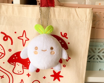 Puede incluir: Un juguete de peluche blanco con forma de rábano con una parte superior verde de hojas. El juguete tiene una cara sonriente con dos pequeños puntos marrones para los ojos y una boca cosida. El juguete está colgado de una bolsa de mano roja con fondo blanco e ilustraciones rojas y blancas.