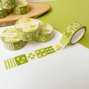 May include: Green daisy blanket washi tape rolls with floral and geometric patterns. The tape features a variety of designs, including daisies, polka dots, and plaid squares. The text "green daisy blanket washi tape" is printed on the tape rolls.