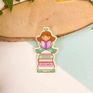 May include: A sticker of a girl sitting on a stack of books. The books are titled "Normal People", "Beach Read", "Pride & Prejudice", "Book Lovers", and "Little Women".