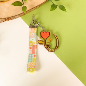 May include: A yellow and pink patterned fabric keychain with a clear acrylic charm featuring a red flower with a yellow stem and leaves.
