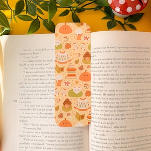 Peut inclure: Un marque-page sur le thème de l'automne avec une variété d'illustrations, notamment des citrouilles, des feuilles, des tasses et un chat. Le marque-page est imprimé sur un fond orange clair avec un motif d'illustrations sur le thème de l'automne. Le texte "cozy" est imprimé dans le coin supérieur droit du marque-page.