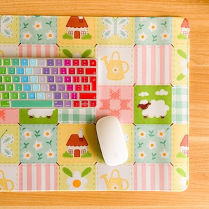 May include: A computer keyboard with rainbow keys, a white mouse, and a patterned mouse mat. The mat features a patchwork design with houses, sheep, butterflies, and flowers in pastel shades, creating a cheerful workspace.