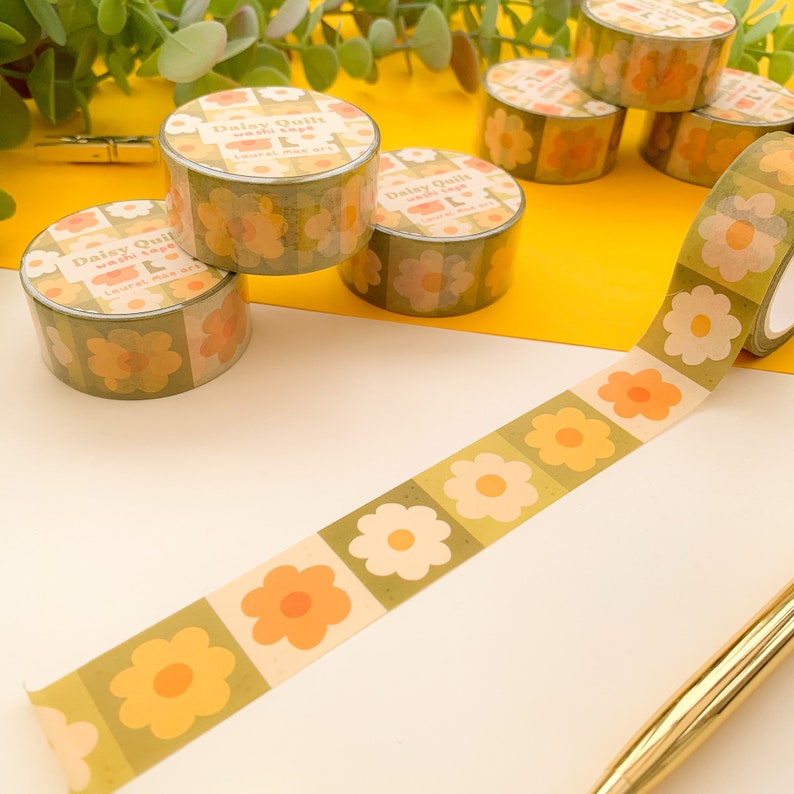 May include: A roll of washi tape with a floral pattern in shades of yellow, orange, and green. The tape features a repeating design of large, stylized flowers with a white center. The tape is labeled "Daisy Quilt Washi Tape Laurel Made Art".