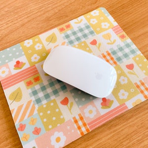 May include: A white wireless computer mouse rests on a rectangular mouse pad featuring a patchwork design of pastel yellow, green, and orange squares. The pattern includes illustrations of small flowers, leaves, gingham checks, and striped motifs.