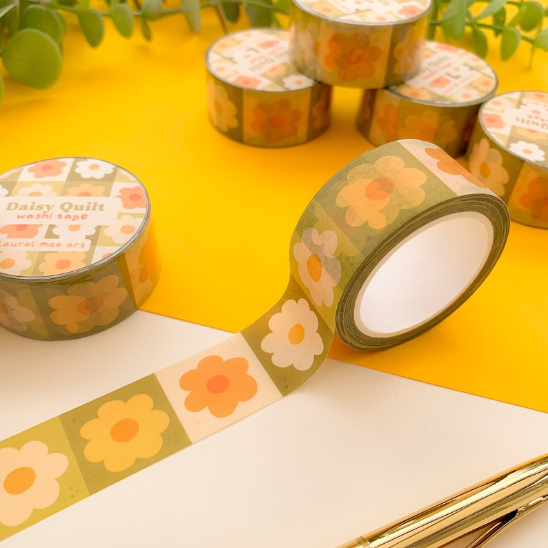 May include: A roll of green and yellow washi tape with a floral pattern. The tape is labeled "Daisy Quilt washi tape Laurel Mae art".