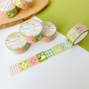 May include: Washi tape rolls with a cottagecore blanket design. The tape displays a repeating pattern of squares with floral, sheep, and butterfly motifs in pastel shades. The text "cottagecore blanket washi tape" is printed on the tape.